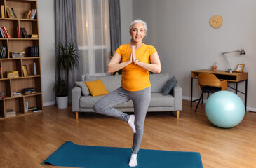 Yoga practice on retirement. Calm senior lady standing in tree pose, keeping balanced, exercising on yoga mat at home - Powered by Adobe
