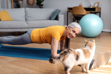 Happy senior woman in sportwear working out at home with cat and watching tutorial lesson on laptop