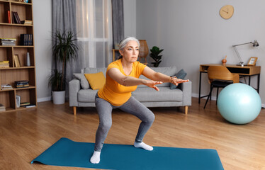 Home workout. Senior lady doing squats on yoga mat, working out at home in living room interior,...
