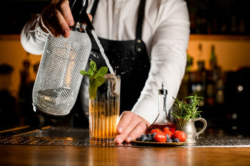 Close-up view of glass with cocktail and sprig of mint in which bartender accurate adds liquid from...
