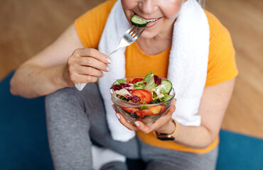 Sports and nutrition concept. Fit senior woman sitting on yoga mat, eating fresh vegetable salad...