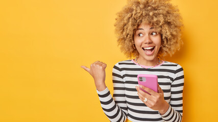 Obraz premium Horizontal shot of cheerful woman holds mobile phone sends text messages in online chat points thumb away dressed in casual striped jumper isolated over yellow background shows announcement.