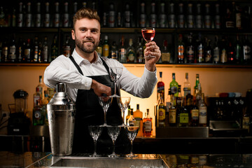 handsome bartender holds glass of drink and large shaker and pyramid of glasses stands nearby on bar