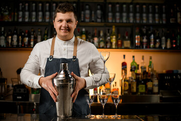 smiling male bartender holds large shaker and pyramid of empty glasses stands nearby on bar