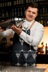 professional bartender holds large shaker and gently pours cocktail on tower of glasses on bar