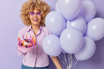 Horizontal shot of positive curly haired woman celebrates birthday in good company holds festive cake and bunch of balloons wears shirt and heart shaped sunglasses isolated over purple background
