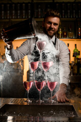Amazing view of the tower made of glasses with cocktail into which bartender pours smoke from bucket