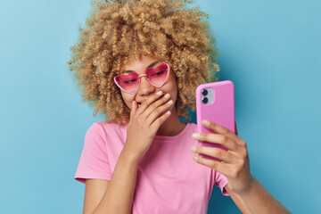 Beautiful woman with curly bushy hair giggles happily covers mouth with hand has video conference uses smartphone dressed in casual pink t shirt and heart shaped sunglasses isolated on blue background