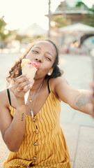 Closeup of smiling young woman with long curly hair with ice cream in her hands making video call on a mobile phone on an urban city background. Close-up of happy girl using smartphone to communicate