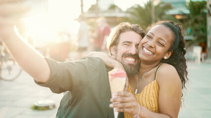 Closeup of smiling interracial couple eating ice cream and taking a selfie on urban city...