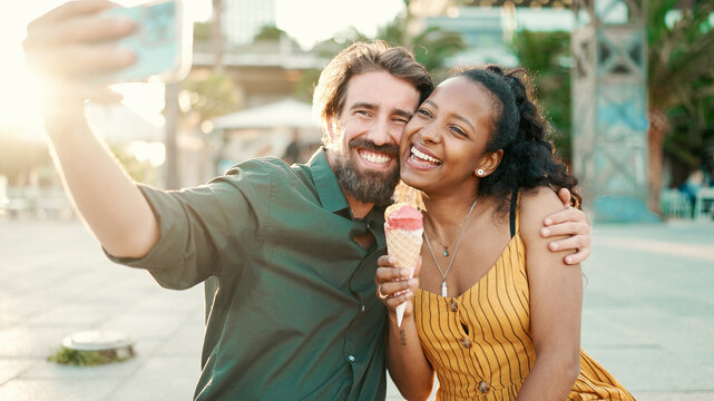 Closeup of smiling interracial couple eating ice cream and taking a selfie on urban city background. Close-up of a man and woman tasting ice cream and video chatting using a mobile phone. Backlight - Powered by Adobe
