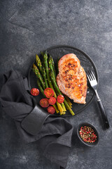 Chicken breast grilled with vegetables. Grilled chicken steak, asparagus and cherry tomatoes in black plate on gray concrete table background. Healthy diet lunch. Barbecue steak fried. Top view.