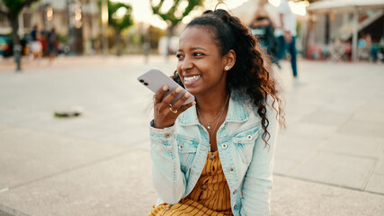 Closeup portrait of young woman with long curly hair using a mobile phone in an urban city background. Close-up of  girl sending a voice message on a smartphone