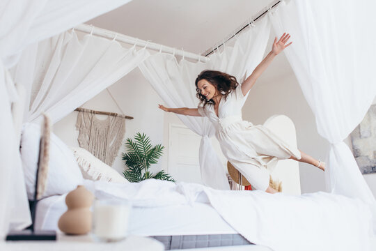 Woman joyfully jumps onto the bed with her arms outstretched as if in flight