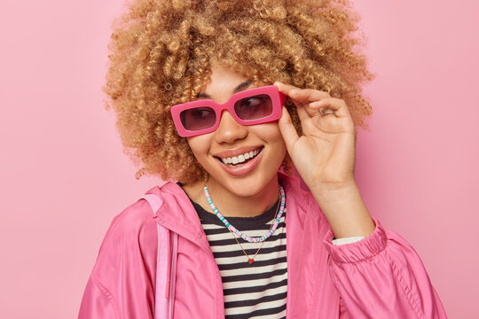 Portrait of cheerful woman with curly bushy hair concentrated away has positive expression wears sunglasses and casual jacket has good mood isolated over pink background. Emotions and style.