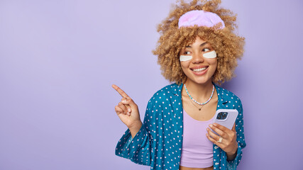 Horizontal shot of cheerful curly haired young woman dressed in nightwear applies beauty patches to reduce puffiness holds mobile phone point left on empty space isolated over purple background
