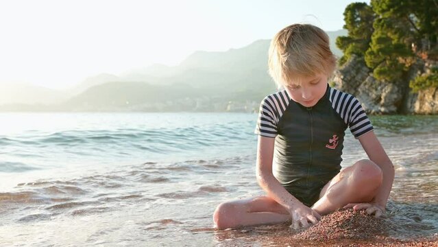 Happy child playing with sand and stones sitting on the seashore. Joyful boy have fun during a beach holiday by the clear warm sea. Leisure for kids and the whole family during school holidays