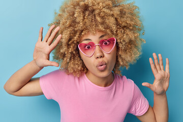 Human facial expressions and reactions concept. Curly haired woman keeps lips folded palms raised looks surprisingly at camera wears trendy pink sunglasses and t shirt isolated over blue background