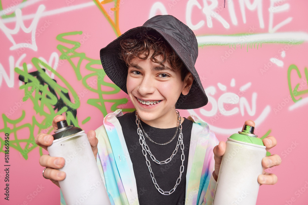 Poster Handsome cheerful hipster boy poses in city against graffiti background wears black panama shirt silver chains braces on teeth holds two cans of paint enjoys favorite hobby. Street art concept