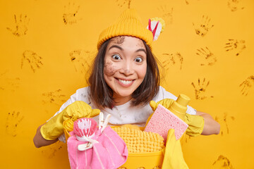 Positive Asian woman with dark hair wears hat and rubber gloves poses with basket full of cleaning...