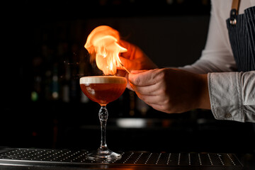 hands of bartender sets fire over glass of cocktail on steel bar counter.