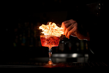 Close-up of crystal glass of cocktail and hands of bartender sets fire over it