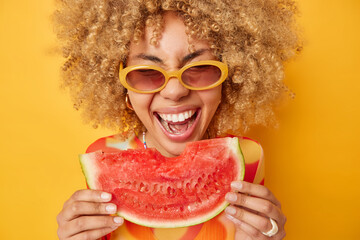 Summer time mood. Positive curly haired European woman eats slice of delicious watermelon holds big...
