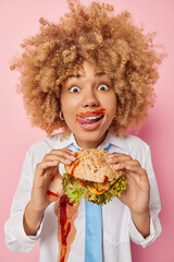 Vertical shot of curly haired woman enjoys eating junk food licks lips smeared with ketchup holds big hamburger has fast food menu. Female student has cheat meal day feels hungry after lectures