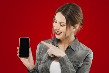 Pretty woman isolated on red background holding phone pointing finger at black copy space screen