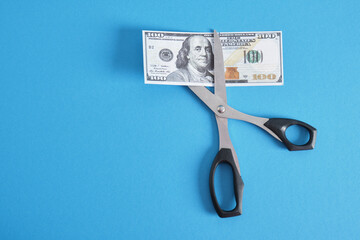 scissors and a bill of 100 dollars on a blue background - obrazy, fototapety, plakaty