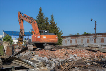 Fototapeta premium Demolition of obsolete dilapidated buildings. A large orange excavator stands on the rubble of a demolished building.