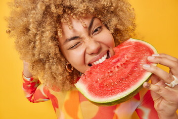 Close up shot of cheerful young European woman with curly hair bites fresh juicy watermelon enjoys eating favorite summer fruit during holidays winks eye poses against vivid yellow background.