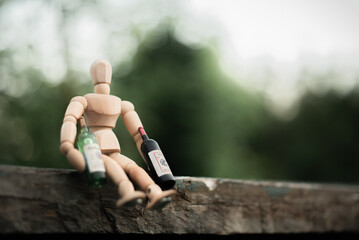 Wooden mannequin doll sitting on a bench with two bottles of vine, like a drunk person. Photo on a natural background with bokeh, horizontal photo