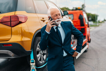 Elegant senior businesman is talking on mobile phone while towing service is helping him with his cars. Roadside assistance concept.