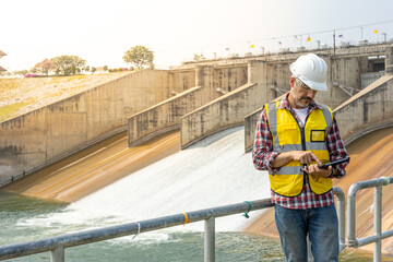 A dam engineering doing his checking routine. He is wearing a white hard hat and yellow transparent vest. He is standing by the rail by the dam.