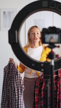 Casual look. Female stylist. Fashion blog. Inspired woman recording two checkered shirts on camera circle led lamp in light home studio interior vertical.