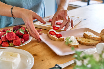 Woman making summer strawberry sandwich. Female hands putting slices of ripe red strawberry on toast with spread stracchino cheese. Healthy eating, fruit dieting brunch.