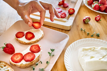 Woman making summer strawberry sandwich. Female hands putting slices of ripe red strawberry on toast with spread stracchino cheese. Healthy eating, fruit dieting brunch.