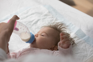Adorable 0-6 baby lying on bed falling asleep sucking milk from bottle, close up shot. Unknown loving caring mother feeding her cute infant daughter before daytime sleep. Nutrition, babyhood concept