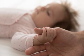 Cute baby falls asleep on bed holds his fathers thumb in hand during carefree healthy sleep, close up cropped shot of dad finger and tiny arm of newborn. Family bond, love, cherish, parenthood concept