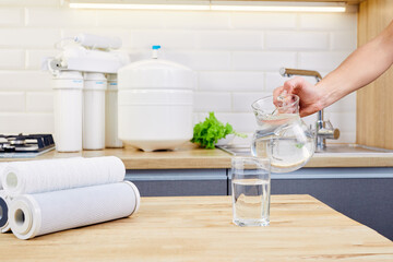 Woman hand pouring purified water from jug into glass in the kitchen. Reverse osmosis filter on...
