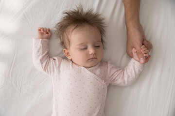 Serene adorable babygirl in pink bodysuit sleeping on bed holding tightly loving daddy thumb, touch...