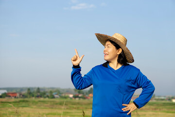 Portrait of Asian woman farmer wears hat, blue shirt, hand on hip, raise finger to point up, Sky background. Copy space for adding text or advertisement. Concept : Agriculture occupation. Happy farmer - Powered by Adobe