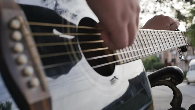 The hand of a teenager boy tunes the strings of an acoustic guitar while sitting on a bench and holding the guitar on his knees. Adjusting the sound of an acoustic guitar, close-up