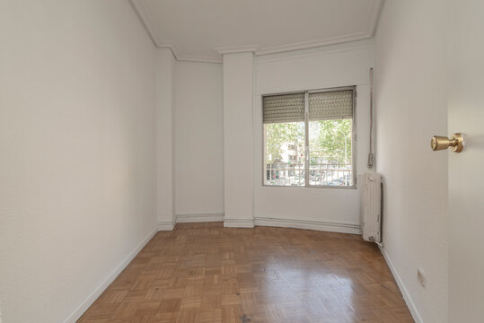 Empty room with oak parquet and white carpentry, oak parquet floor and cast iron radiator and plain white painted walls with aluminum and glass window overlooking the street with trees