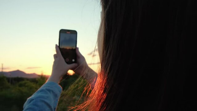 Selective focus of cute young caucasian woman 20s using smartphone, record video or make photos of amazing sunset and landscape scenery. Concept adventure travel blogger. Outdoors living lifestyle