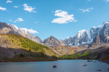 Pure azure alpine lake with view to forest hill and giant snow mountains in autumn sunny day. Glacial lake against huge snow covered mountain range in bright sun. Vivid autumn colors in high mountains