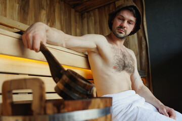 Serious man resting in a sauna and draws water from a bucket with a ladle