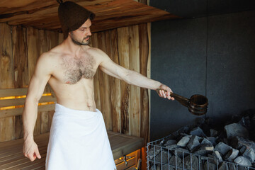 Muscular handsome man pours water on hot stones in the sauna
