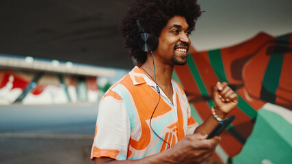 Close-up of cheerful young African American man wearing shirt listening to music in headphones and dancing on urban city background. Lifestyle concept.